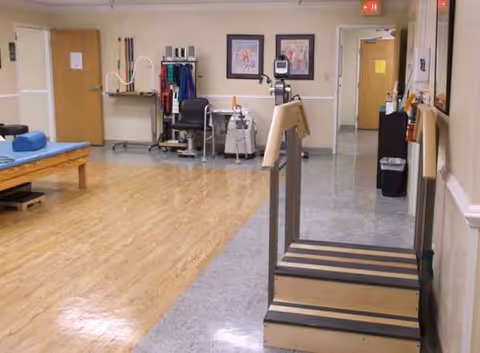 Therapy room with wooden floor, gait training steps, treatment table, and medical equipment.