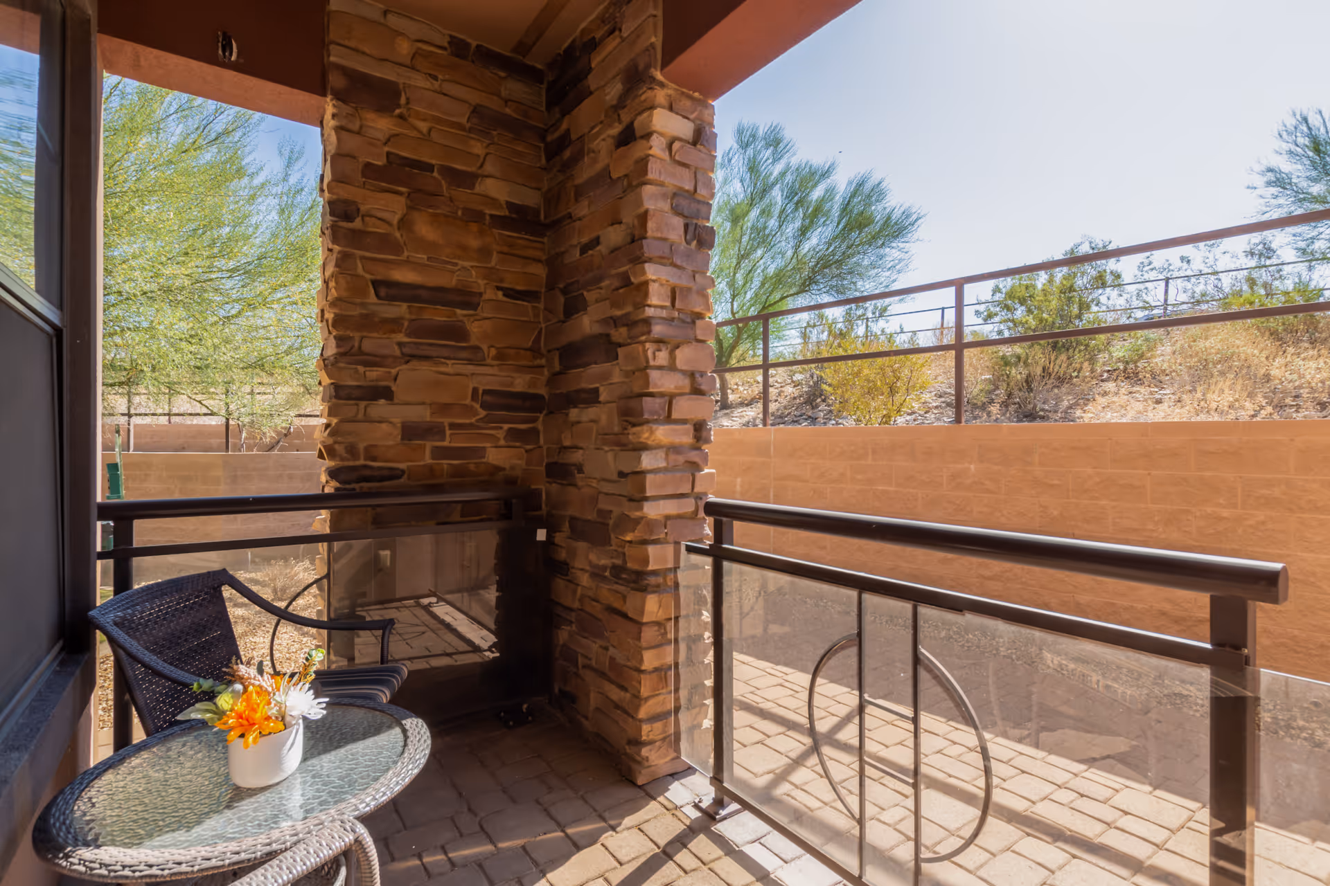 Small outdoor patio area with a glass-top round table holding a small flower arrangement, a single wicker chair, stone pillar walls, and a metal railing overlooking a dry landscape with trees and bushes.