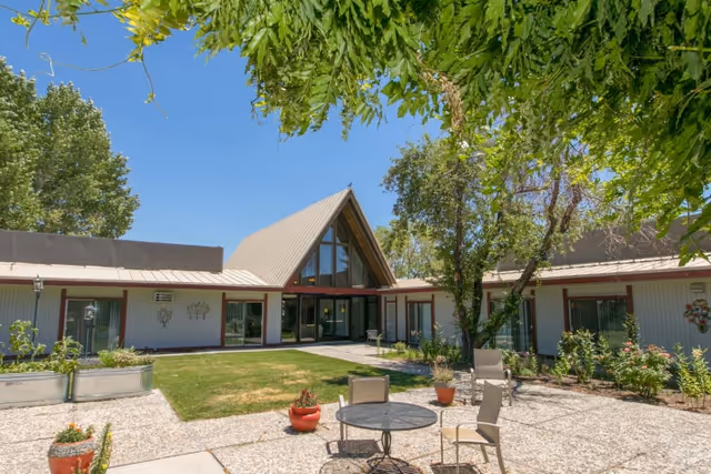 Courtyard of a rehabilitation center with a triangular glass entrance flanked by single-story wings, patio seating, potted plants and trees under a clear blue sky.