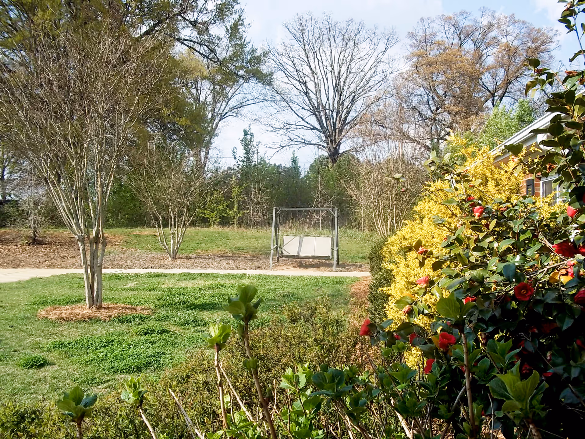 Outdoor garden area with green grass, trees, bushes with red flowers, and a metal swing bench. A building with yellow siding is partially visible on the right side.