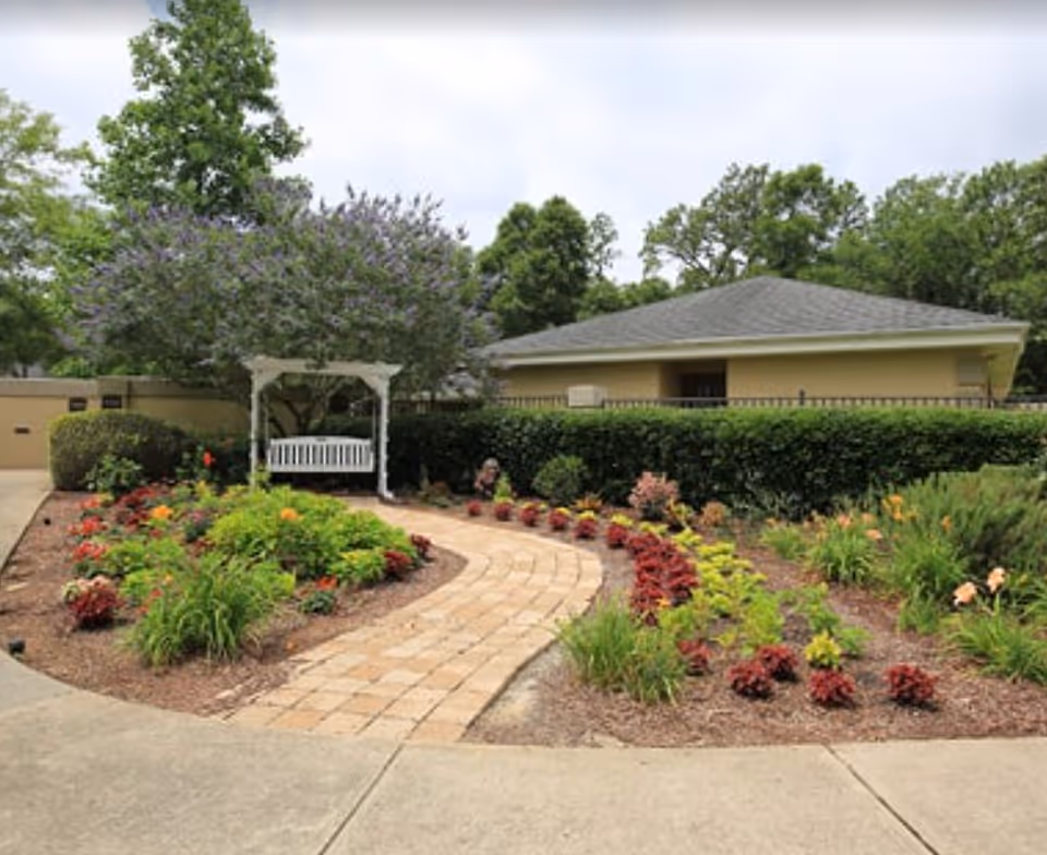 Curved paved walkway through a landscaped garden leading to a white arbor with a bench in front of a single-story building.