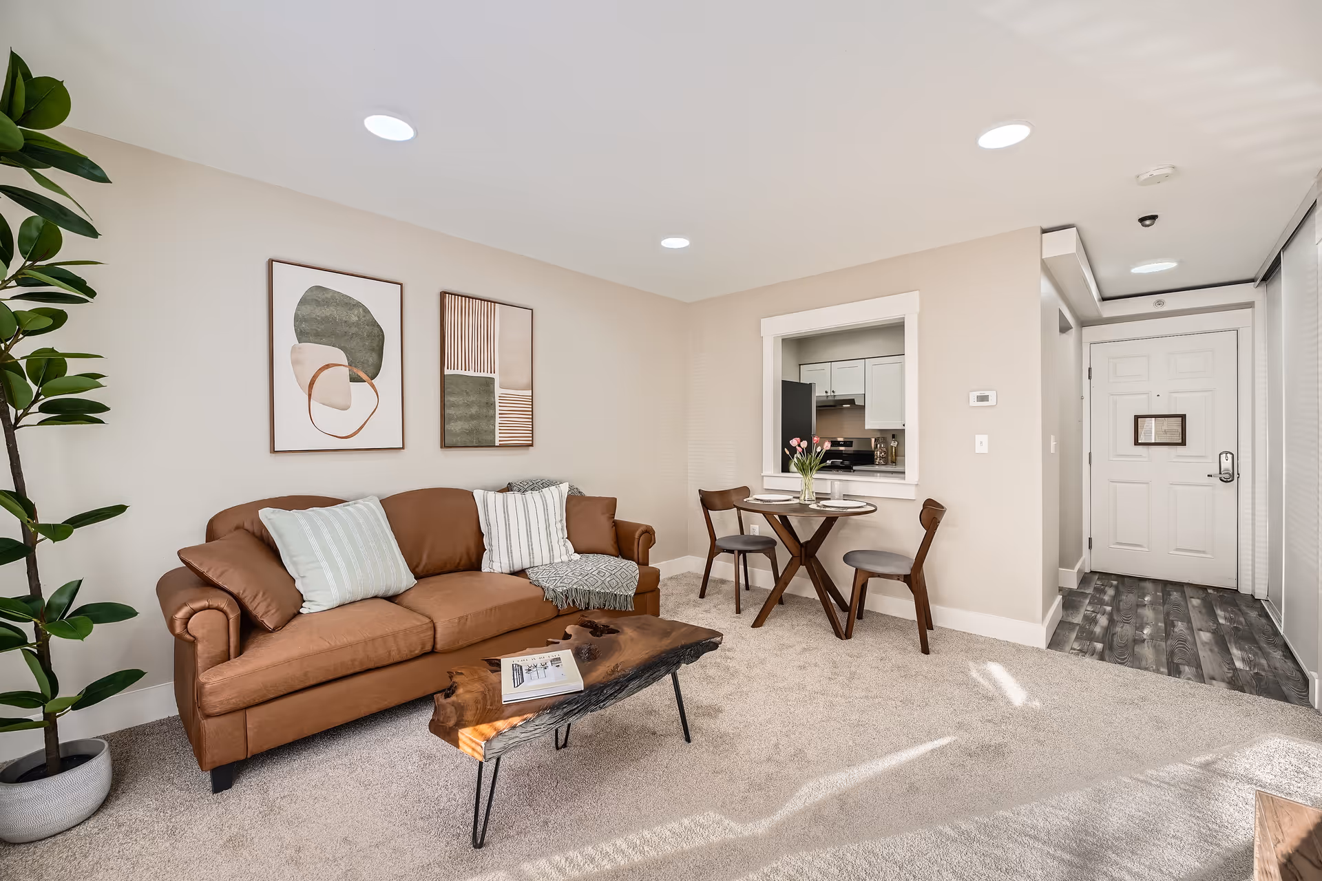 A cozy living room with a brown leather sofa adorned with pillows and a throw blanket. A rustic wooden coffee table with a book on it sits in front of the sofa. Two abstract art pieces hang on the beige wall above the sofa. To the right, there is a small round dining table with two chairs and a vase of pink tulips. A pass-through window reveals a glimpse of a white kitchen. The entrance door and hallway with wood flooring are visible in the background. A tall green plant is placed in the corner on the left side.