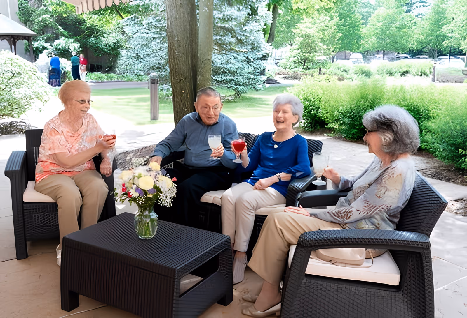 Four elderly individuals sitting outdoors on black wicker chairs around a matching table with a vase of flowers, enjoying drinks and engaging in conversation in a garden-like setting with greenery and trees in the background.