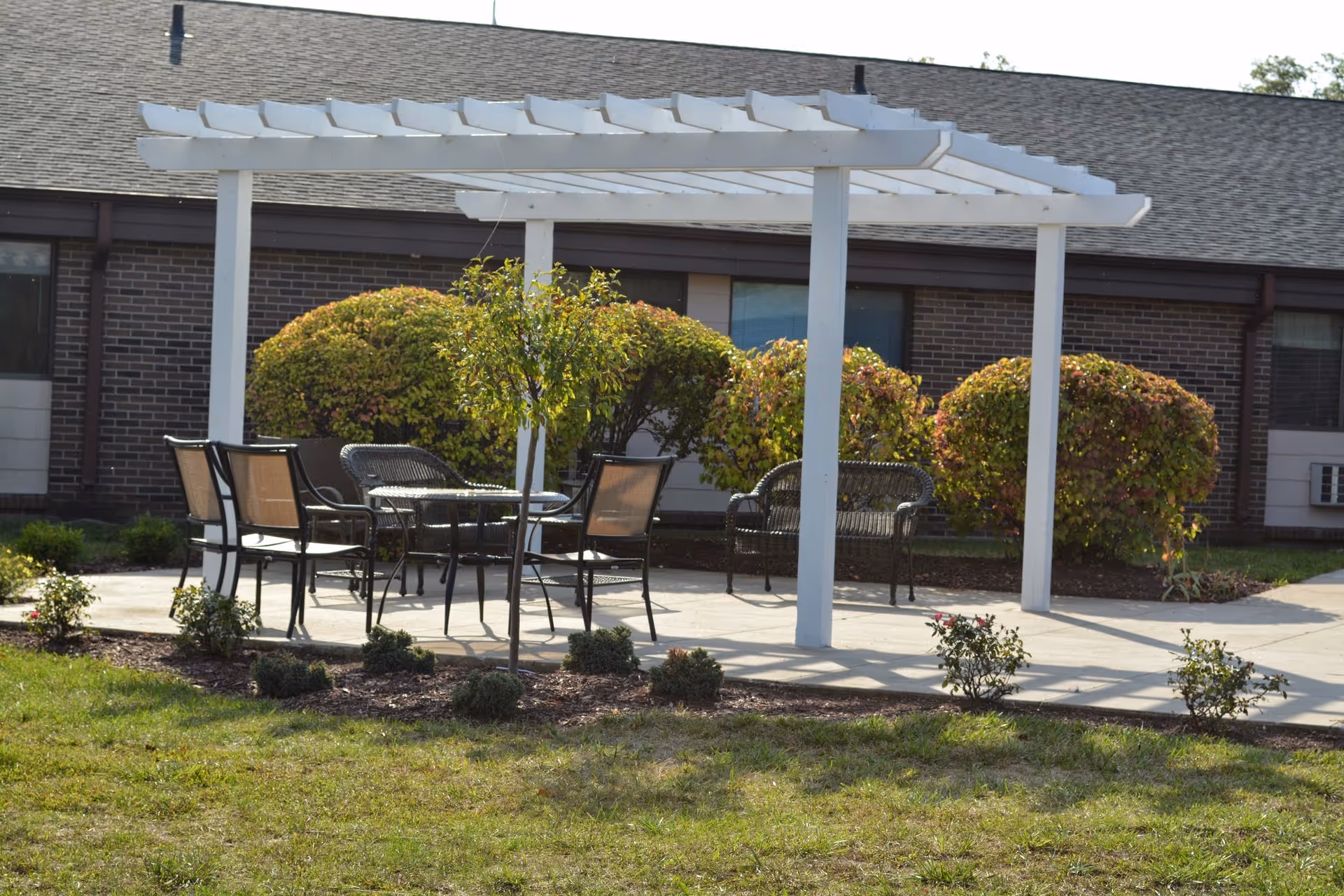 Outdoor seating area with a white pergola, metal chairs, and a table on a concrete patio surrounded by small bushes and grass, with a brick building in the background.