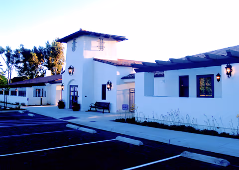 Exterior view of a white building with Spanish-style architecture, featuring a tower-like structure, outdoor wall lanterns, a bench near the entrance, and an empty parking lot in the foreground.