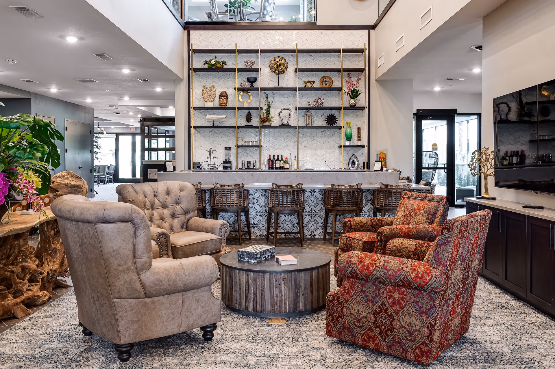 A cozy living area with two beige armchairs and two patterned red armchairs arranged around a round wooden coffee table. Behind the seating area is a bar with five wooden bar stools and decorative shelves displaying various ornaments and bottles. The space is well-lit with recessed ceiling lights and has a large flat-screen TV mounted on the right wall.