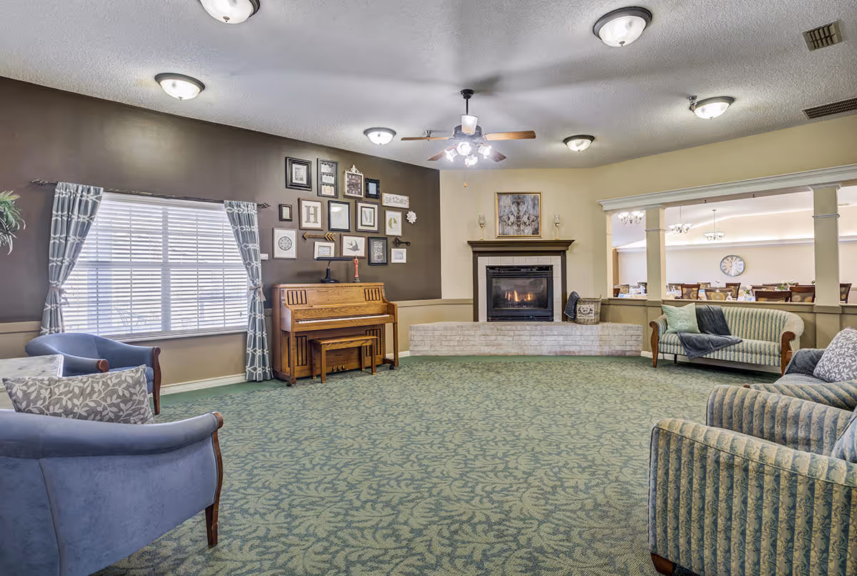 A spacious living room with patterned green carpet, a ceiling fan with lights, and multiple ceiling light fixtures. The room features a fireplace with a painting above it, a wooden piano against a dark accent wall decorated with framed pictures and the word 'Home'. There are several upholstered chairs and sofas arranged around the room, and a large window with patterned curtains letting in natural light. Through an open partition, a dining area with tables and chairs is visible.