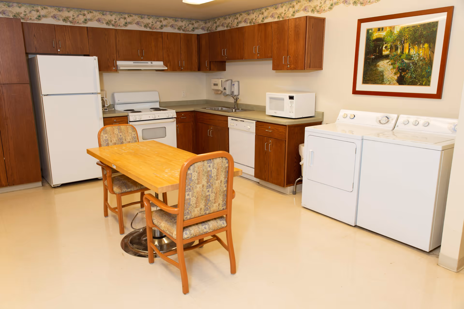 A kitchen area in a senior living facility featuring wooden cabinets, a white refrigerator, stove, dishwasher, microwave, and a washer and dryer set. There is a wooden table with two cushioned chairs in the center of the room. A framed painting hangs on the wall above the washer and dryer. The room has a light-colored floor and a floral wallpaper border near the ceiling.