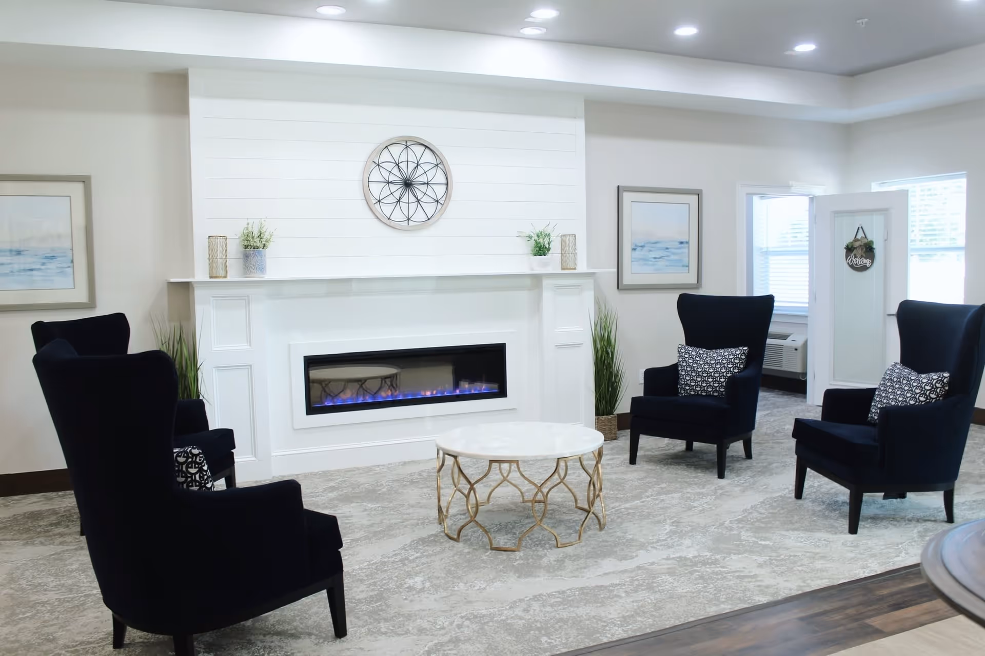 A cozy living room area with four dark blue armchairs arranged around a round white marble coffee table with a gold metal base. The room features a modern electric fireplace set in a white mantel with decorative plants and a circular wall decor above it. Two framed paintings hang on the walls, and there is a white folding screen with a 'Welcome' sign near a window with blinds.