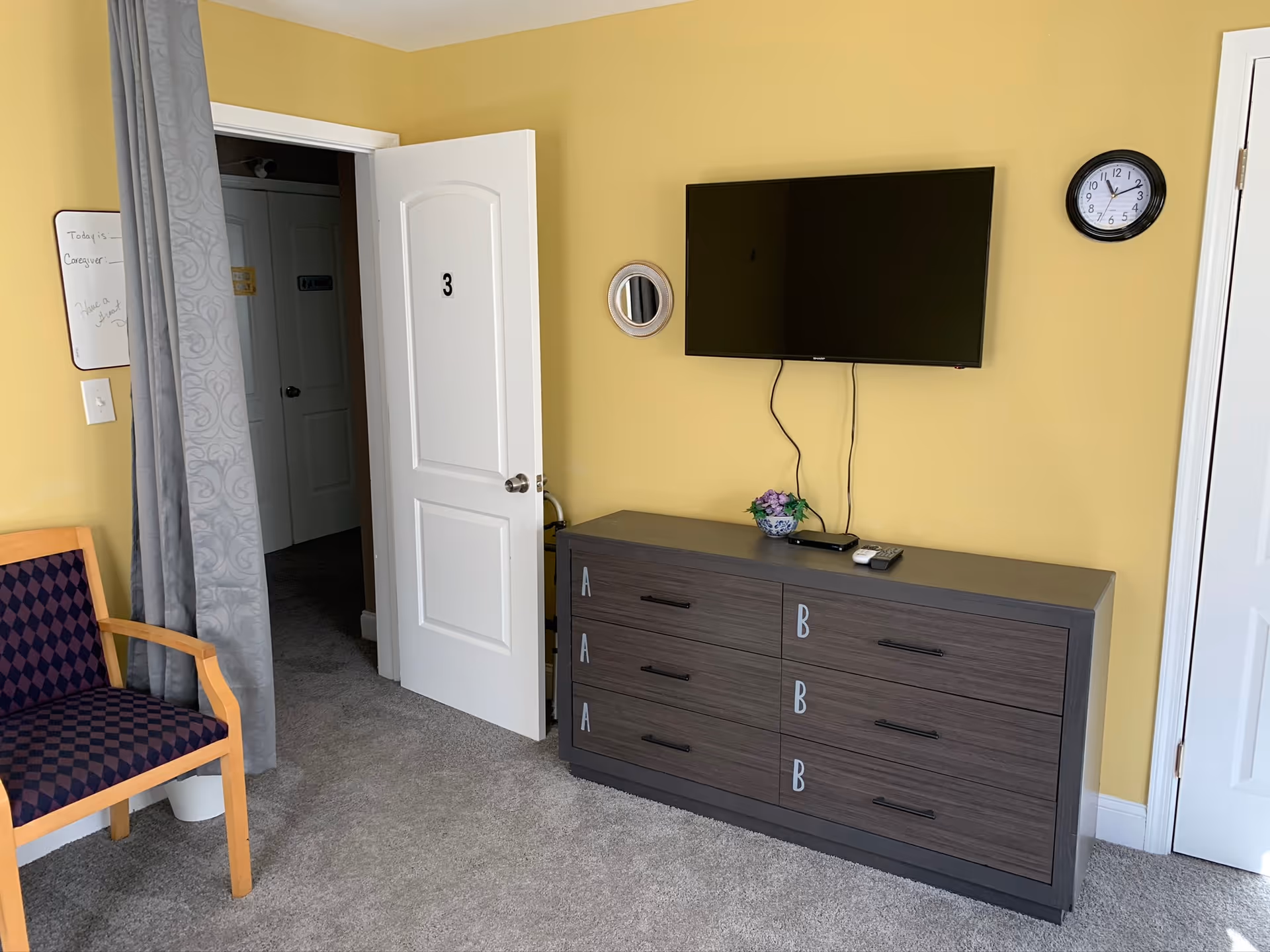 A room with yellow walls featuring a wall-mounted flat screen TV above a dark wooden dresser with labeled drawers. A small round mirror and a clock are mounted on the wall. There is a wooden chair with a purple patterned cushion, a gray curtain, and an open white door marked with the number 3 leading to a hallway.