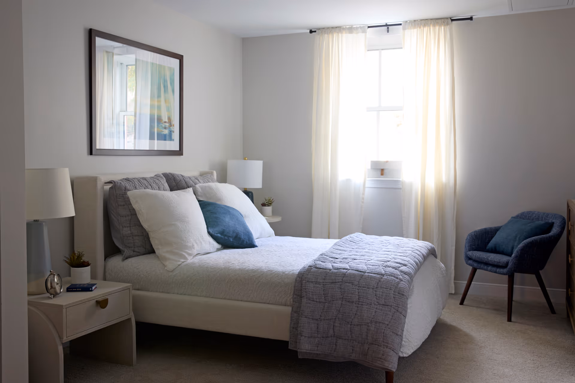 A neatly made bed with white and gray bedding and a blue accent pillow in a bright bedroom. There is a nightstand with a lamp, a small plant, and an alarm clock on the left side of the bed. A framed picture hangs on the wall above the bed. A window with sheer white curtains lets in natural light. A blue upholstered chair with a matching pillow is positioned near the window on the right side of the room.
