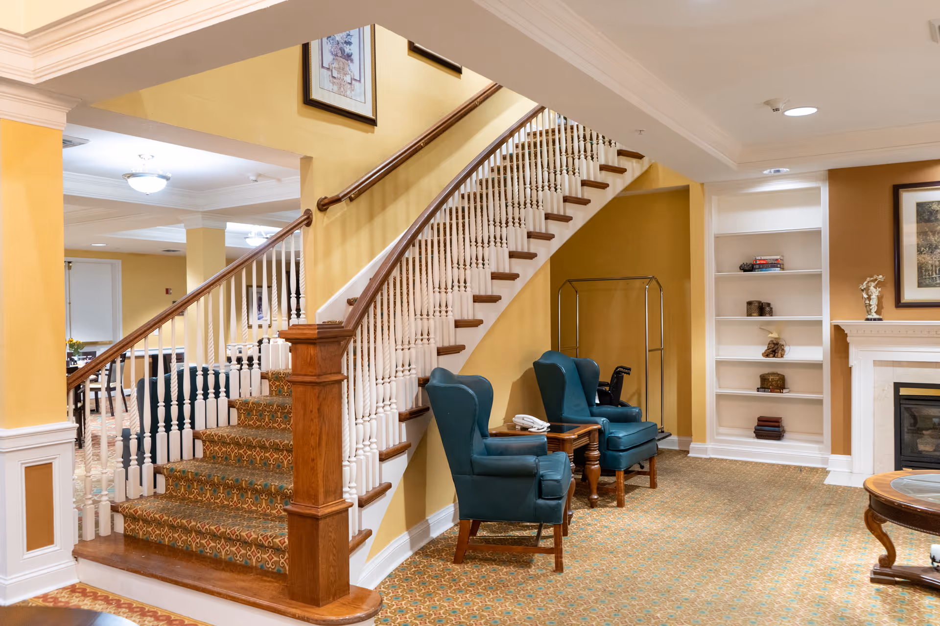 A bright senior living facility interior showing a curved staircase, two teal armchairs, built-in shelving, and a fireplace.