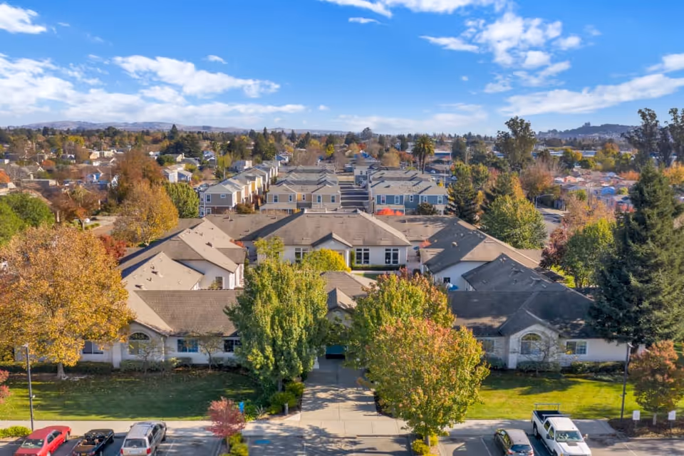 Aerial view of a single-story memory care complex surrounded by trees, parking spaces, and nearby houses under a blue sky.