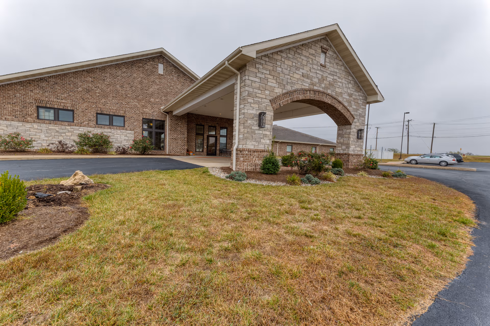 Exterior view of Cedarhurst Senior Living of Waterloo showing the building entrance with a covered archway, brick and stone facade, landscaped grass and shrubs, and a parking area with cars in the background under a cloudy sky.