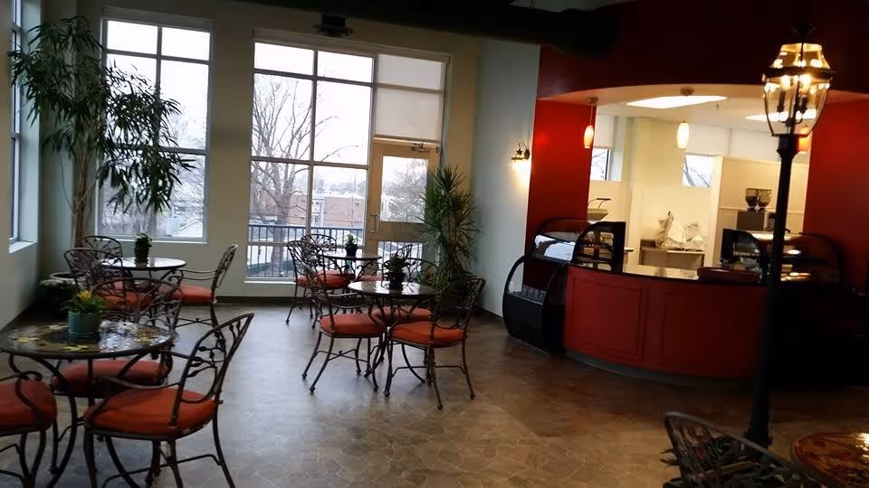 Interior view of a dining area with several round tables and wrought iron chairs with red cushions. Large windows and a glass door provide natural light and a view of trees outside. There is a red counter area with display cases and pendant lights, and a tall lamp post-style light fixture near the seating area. Potted plants are placed around the room.