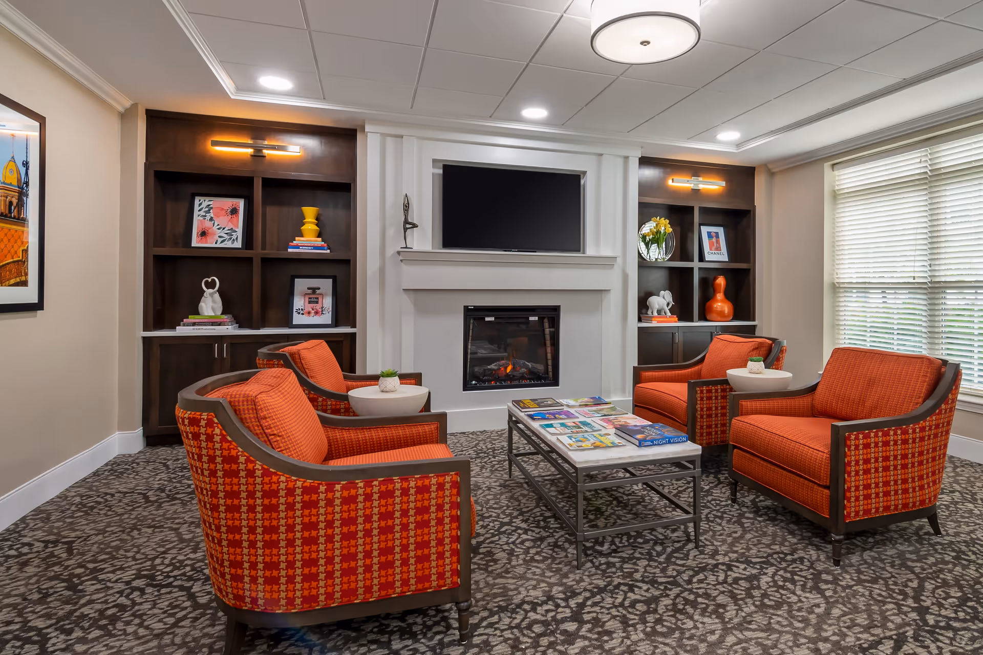 Cozy common lounge with four orange armchairs around a coffee table facing a fireplace and wall-mounted TV, flanked by built-in shelving.