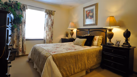 Well-lit furnished bedroom with a bed, nightstands, lamps, framed art, and a window with floral curtains.