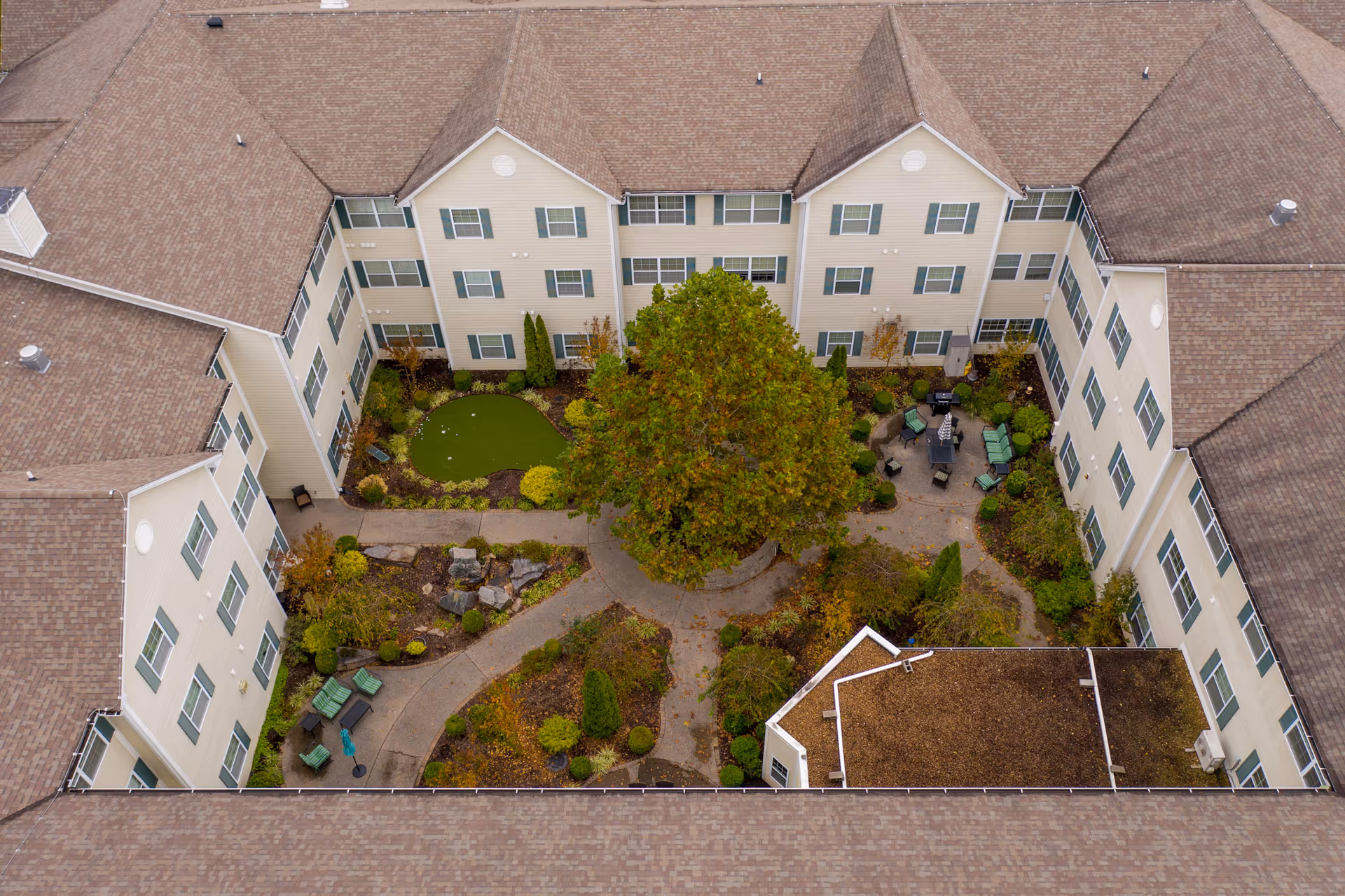 Aerial view of a courtyard garden within a senior living facility. The courtyard features a large tree in the center, surrounded by walking paths, landscaped bushes, a small putting green, and seating areas with chairs and tables. The building surrounding the courtyard has beige siding with green window shutters and a brown shingled roof.