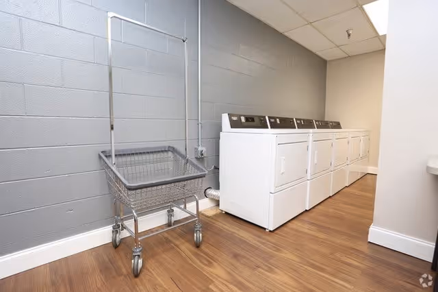Laundry room with a row of white washing machines and dryers along the right wall, a metal laundry cart with wheels on the left side, and wood-look flooring with gray painted cinder block walls.