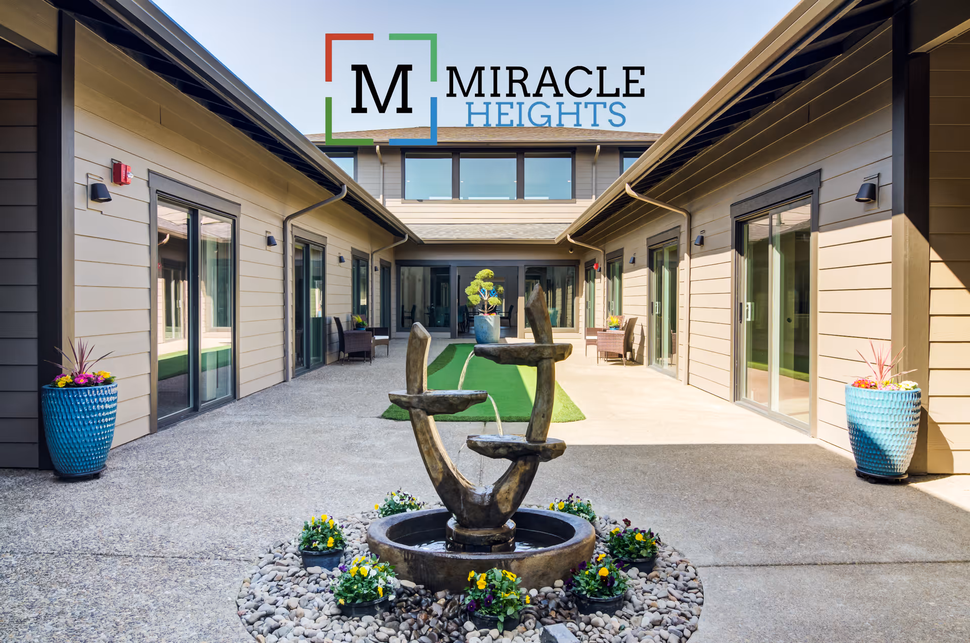 Outdoor courtyard area of Miracle Heights facility featuring a modern water fountain surrounded by small flowers and stones, with beige buildings on both sides having large glass sliding doors and blue planters with flowers near the entrances.