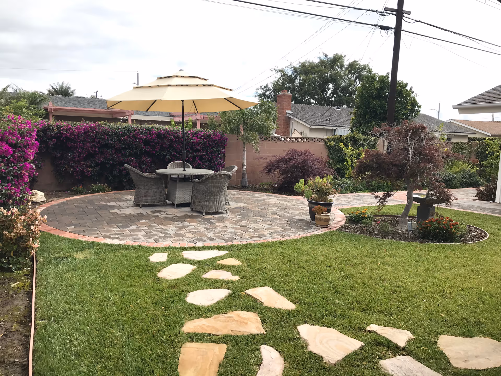 A well-maintained outdoor garden area with a stone pathway leading to a circular paved patio. The patio has a round table with four wicker chairs and a large beige umbrella. Surrounding the patio are various plants, bushes, and small trees, with a residential neighborhood visible in the background.