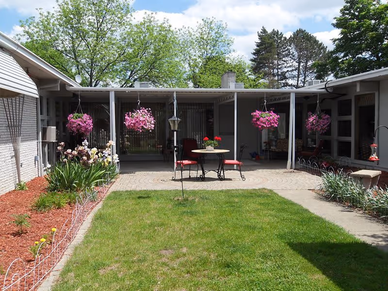 Sunny courtyard of a single-story senior living facility with a small lawn, patio table and chairs, hanging flower baskets and a covered walkway.