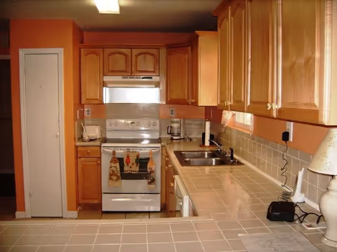 A kitchen with wooden cabinets, a white stove with oven mitts hanging on the handle, a white toaster, a coffee maker, a double sink, and a tiled countertop. The walls are painted orange and there is a lamp on the right side of the counter.