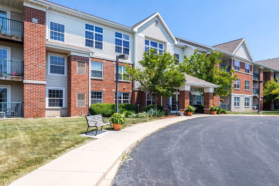 Exterior view of a senior living facility named Killarney Kourt showing a three-story building with red brick and white siding. There is a covered entrance with potted plants and trees, a bench on the sidewalk, and a paved driveway in front under a clear blue sky.