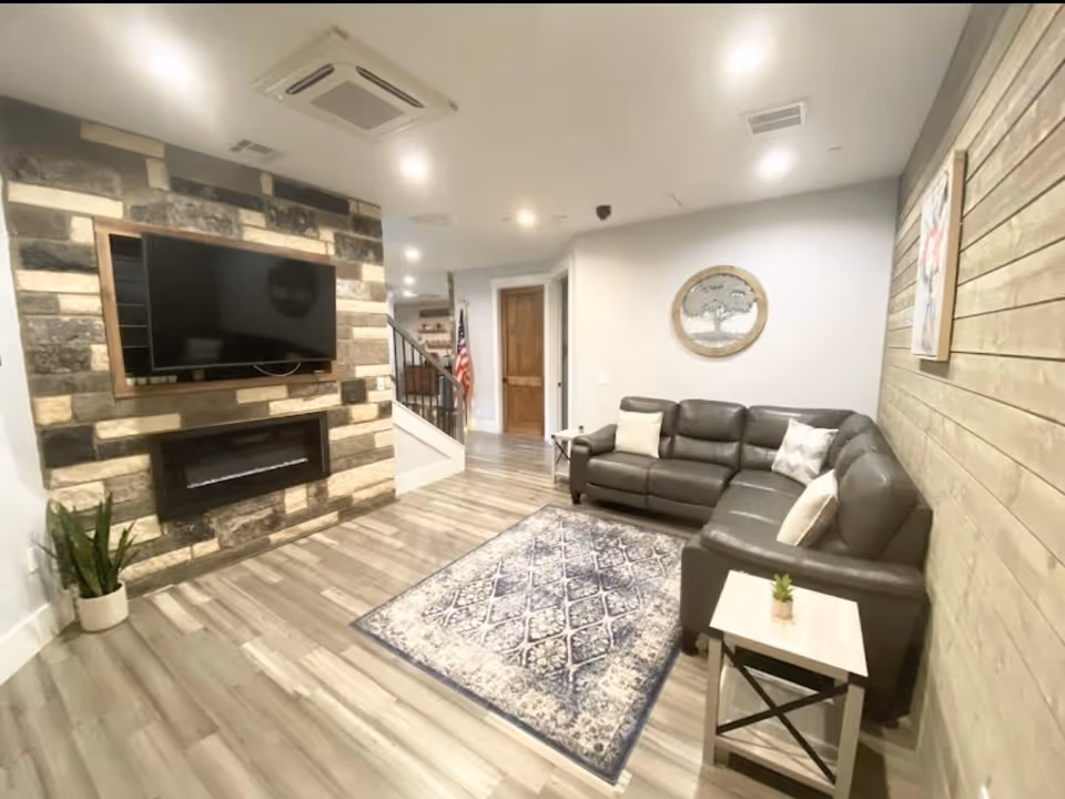 A cozy living room with a dark brown leather sectional sofa adorned with light-colored pillows, a patterned area rug on light wood flooring, a stone accent wall with a mounted flat-screen TV and an electric fireplace below it, a small side table with a plant, and wooden paneling on the adjacent wall. The room is well-lit with recessed ceiling lights and has a staircase and an American flag visible in the background.