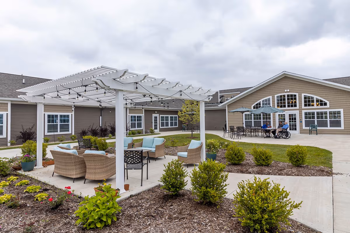 Outdoor courtyard area of Riley's Grove Assisted Living & Memory Care featuring a white pergola with string lights, wicker seating with light blue cushions, landscaped garden beds with shrubs and flowers, and a patio with tables, chairs, and umbrellas where three people are seated, one in a wheelchair.