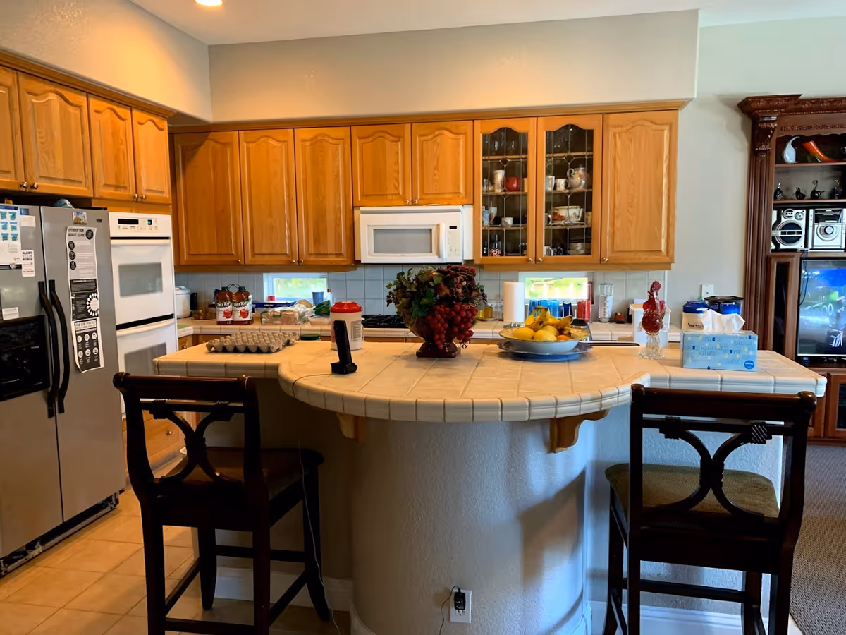 A kitchen with wooden cabinets, a white microwave, double oven, and stainless steel refrigerator. There is a curved tile countertop island with two wooden chairs. The countertop has a fruit bowl, a decorative plant, a tissue box, and other kitchen items. In the background, there is a wooden entertainment center with a TV and stereo system.