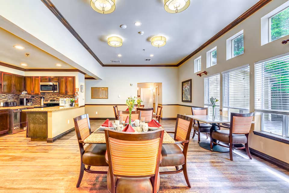 Bright dining area with wooden tables and chairs, set with red napkins and white mugs. Large windows with blinds let in natural light. The room features a kitchen area with dark wood cabinets and stainless steel appliances in the background.