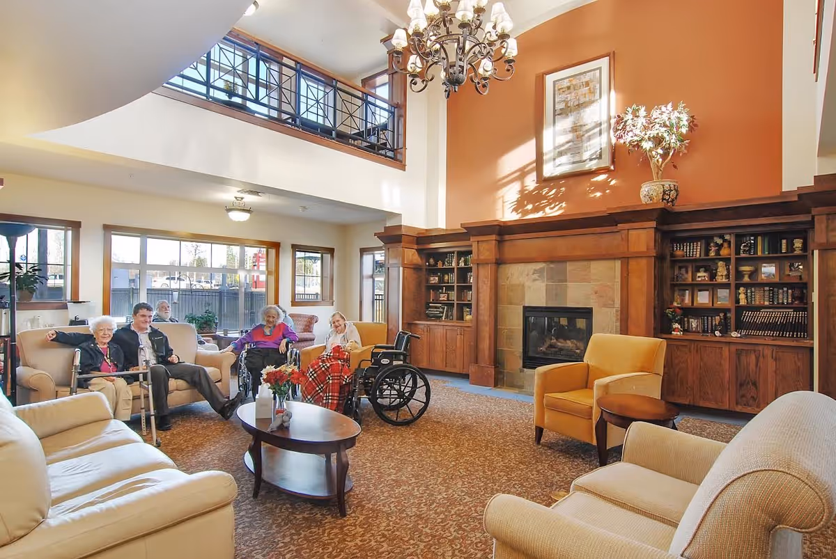 A spacious and well-lit living room in a senior living facility with a high ceiling and large windows. The room features a fireplace with wooden built-in bookshelves on either side, a chandelier, and various comfortable seating options including armchairs, sofas, and wheelchairs. Five elderly people are seated and smiling, enjoying the cozy atmosphere.