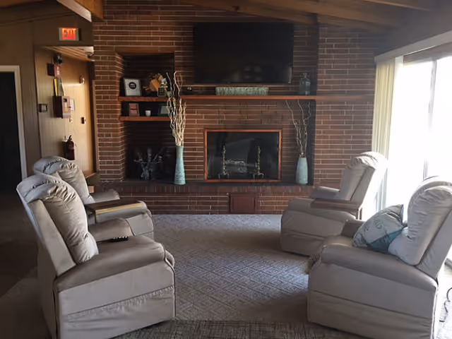 A cozy living room with four beige recliner chairs arranged around a brick fireplace. Above the fireplace is a flat-screen TV and a decorative 'WELCOME' sign. The room has a carpeted floor and large windows with vertical blinds letting in natural light.