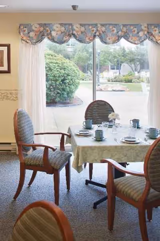A dining room with a table set for meals, surrounded by chairs, near a large window.