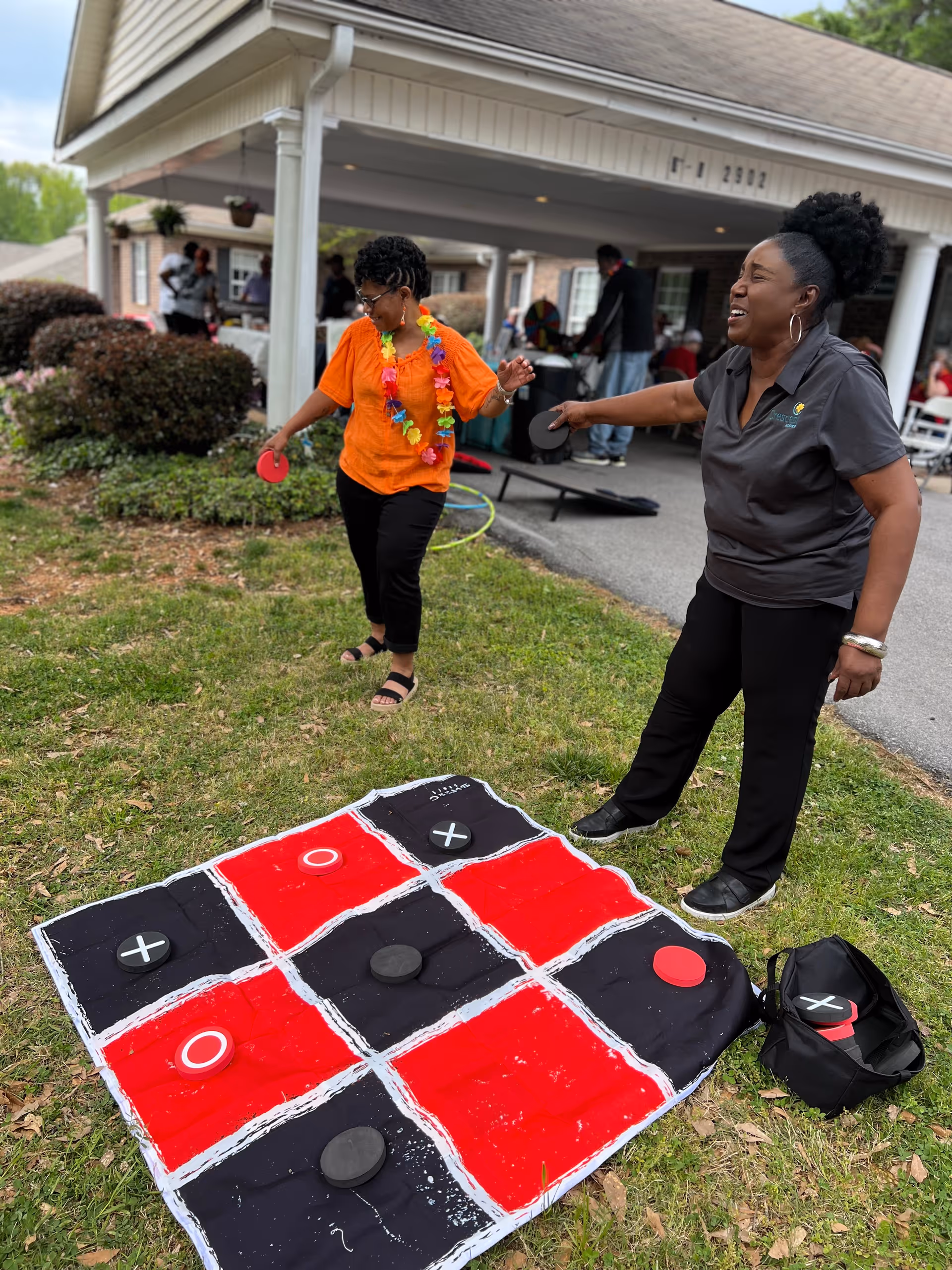 Two women playing a large outdoor tic-tac-toe game on a red and black mat on the grass outside a building with a covered porch. One woman is wearing an orange shirt and a colorful lei, and the other woman is wearing a dark gray shirt and black pants. Other people and outdoor games are visible in the background.