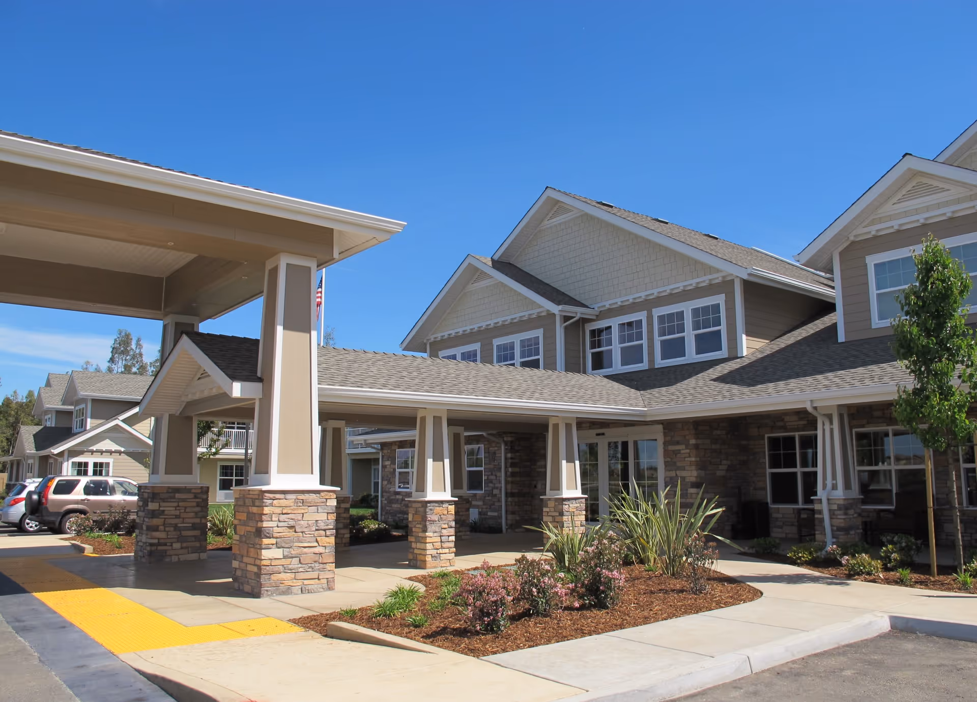 Exterior view of Camden Springs Gracious Retirement Living facility showing a covered entrance with stone pillars, landscaped garden beds with plants and flowers, and multiple windows under a clear blue sky.