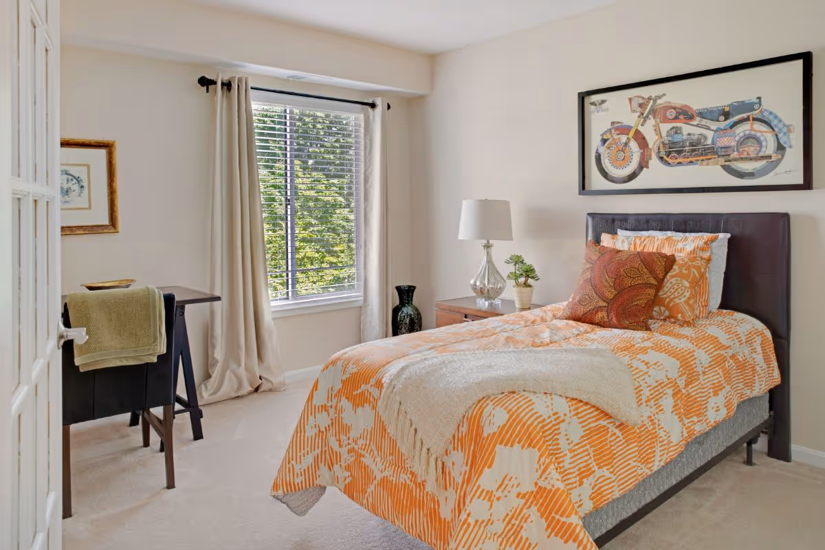 A bright bedroom with a single bed covered in an orange and white patterned bedspread, accented with decorative pillows and a beige throw blanket. Next to the bed is a wooden nightstand with a silver lamp and a small potted plant. A framed picture of a motorcycle hangs on the wall above the bed. A window with beige curtains and white blinds lets in natural light, and a small desk with a chair and a green towel draped over the back is positioned near the window.