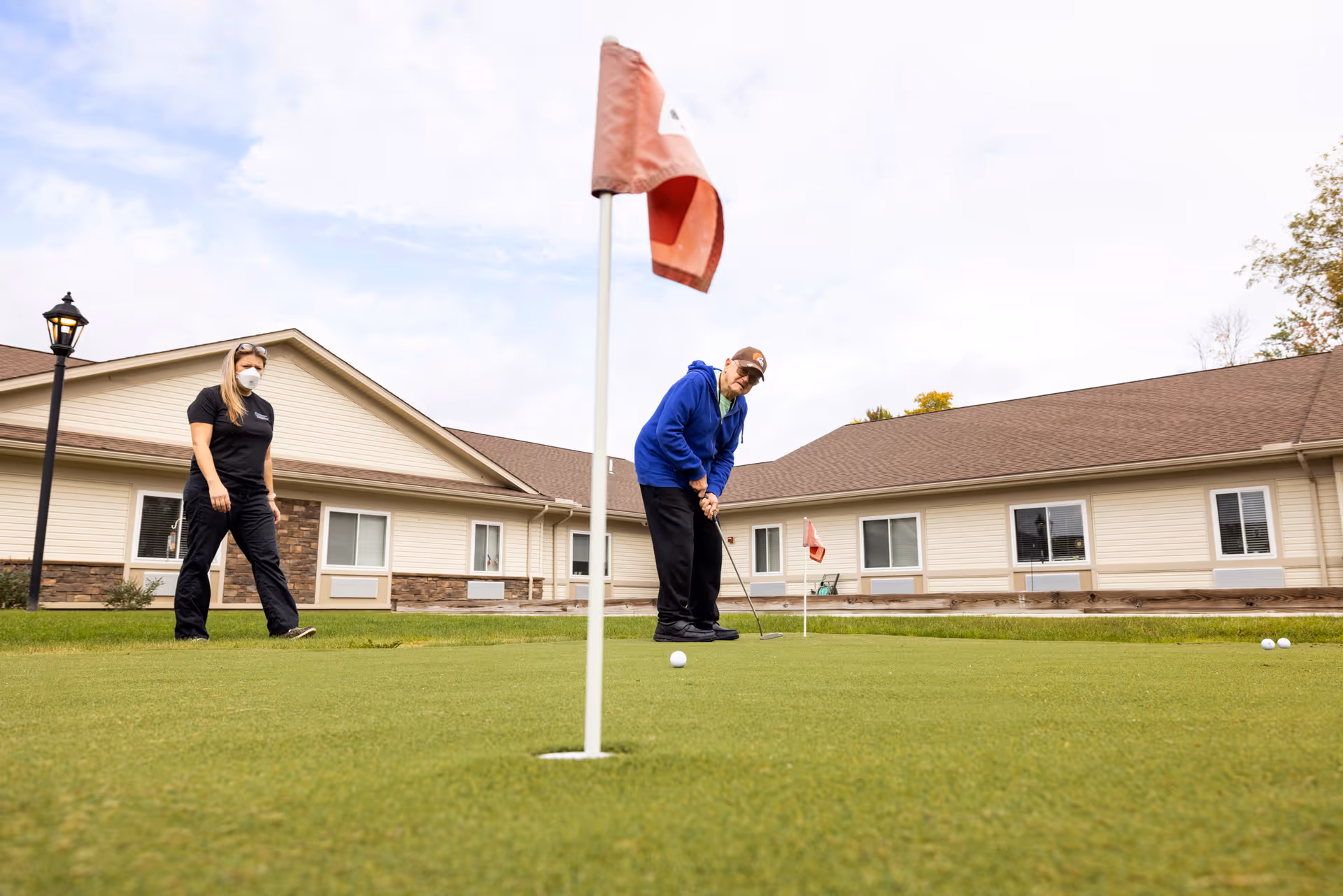 An elderly man wearing a blue jacket and cap is putting on a golf green while a woman in black scrubs and a face mask watches nearby. They are outside near a single-story building with beige siding and brown roof, under a partly cloudy sky.