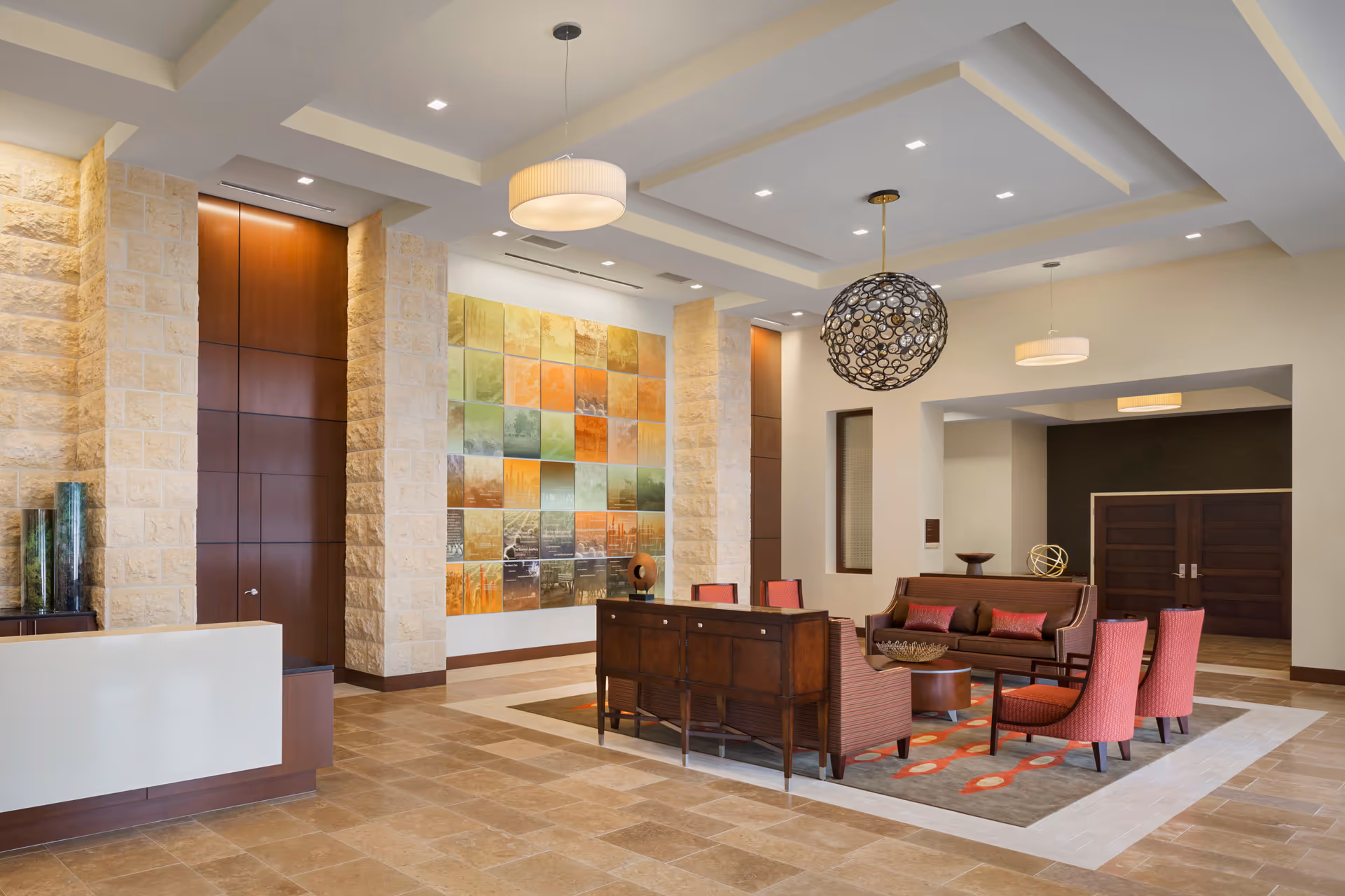 A spacious and modern senior living facility lobby with beige tiled floors, stone accent walls, and a high ceiling with recessed lighting. The seating area includes a brown sofa, four red upholstered chairs, and a wooden console table on a patterned area rug. Two pendant lights hang from the ceiling, one round and decorative, the other simple and white. A colorful wall art installation is displayed on one wall, and there are double wooden doors in the background.