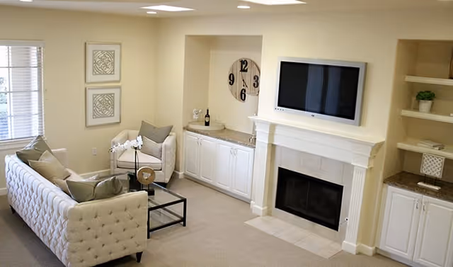 A cozy living room with a white tufted sofa, a beige armchair, a glass coffee table with a decorative orchid, a wall-mounted flat screen TV above a white fireplace, built-in white cabinets with granite countertops, and a large decorative wall clock.