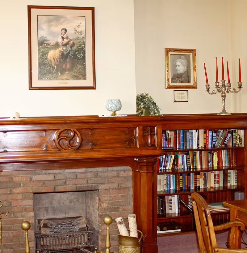 A cozy interior room featuring a wooden fireplace mantel with decorative carvings, a brick fireplace with firewood and brass andirons, a bookshelf filled with books, a wooden chair, and framed pictures on the beige walls. A silver candelabra with red candles and a small plant are placed on the mantel.