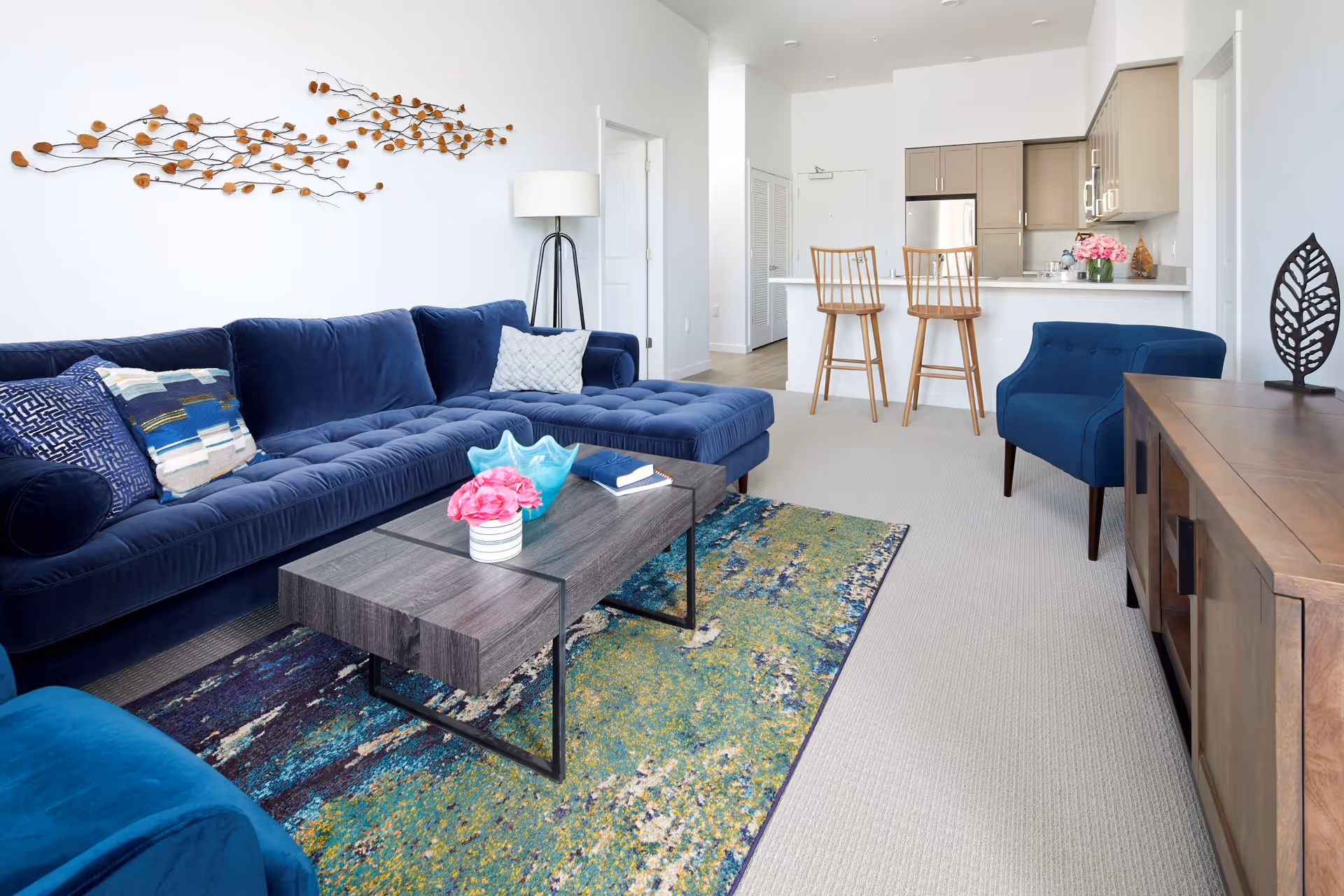 Bright and modern living room with a navy blue sectional sofa adorned with patterned pillows, a wooden coffee table with decorative items including a pink flower and a blue glass bowl, a colorful area rug, a blue armchair, and a wooden sideboard with a leaf-shaped sculpture. In the background, there is a kitchen area with two wooden bar stools at a counter, beige cabinets, and stainless steel appliances.