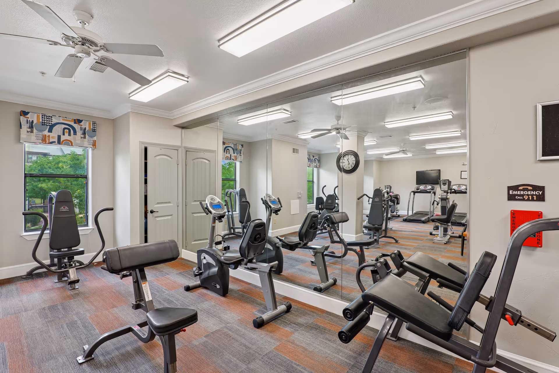 A well-lit fitness room with various exercise equipment including stationary bikes, weight machines, and treadmills. The room has large mirrors on one wall, a ceiling fan, windows with patterned valances, and a clock mounted on a pillar. There is an emergency call sign on the wall.