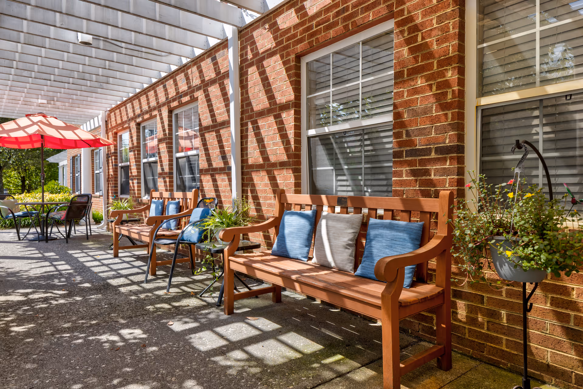 Outdoor patio area with wooden benches adorned with blue and gray cushions, a hanging plant, and a table with chairs under a red umbrella. The area is shaded by a white pergola attached to a brick building with multiple windows.