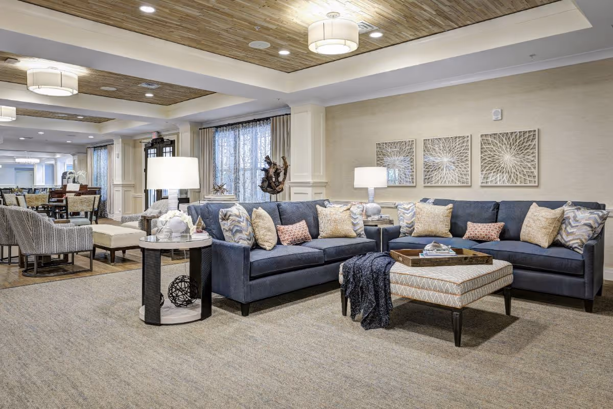 A spacious and well-lit living room area in a senior living facility featuring two blue sofas with multiple patterned cushions, a patterned ottoman with a tray on top, two side tables with lamps, and decorative wall art. In the background, there is a dining area with tables and chairs, and large windows with curtains.