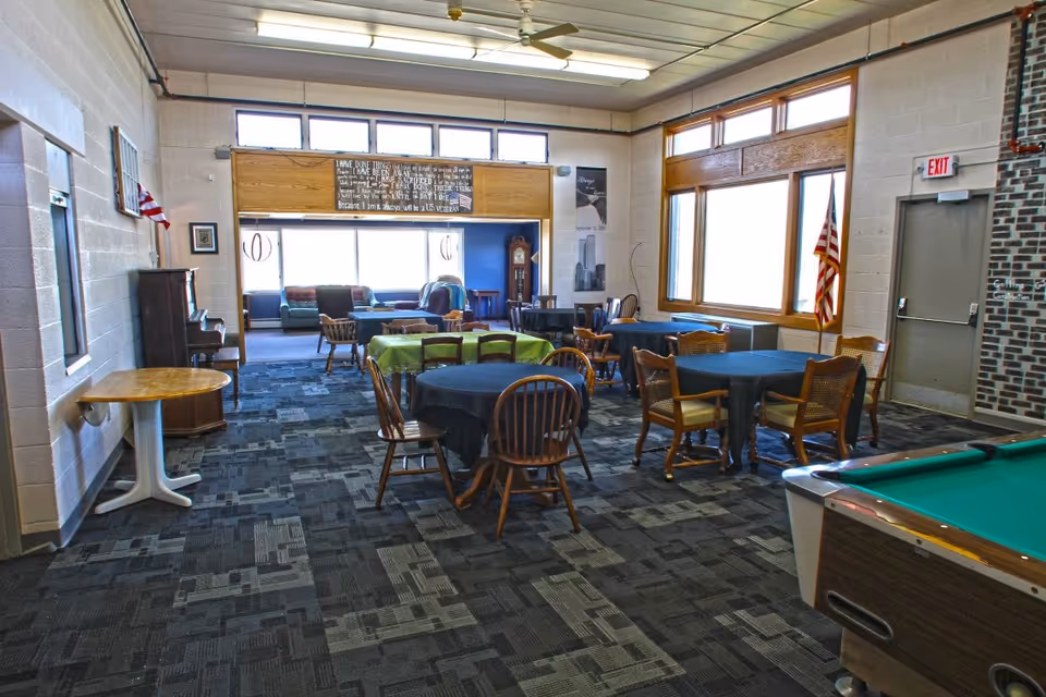 Large multipurpose senior community room with round tables and chairs, a pool table, and a seating area near windows.