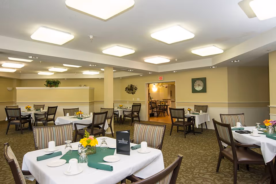 A dining room with multiple tables covered in white tablecloths and green runners, each set with plates, cups, glasses, and napkins. The room has beige walls, patterned carpet, and ceiling lights. There are chairs around each table and floral centerpieces on the tables.