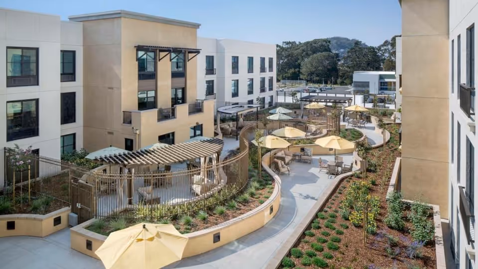 Outdoor courtyard area of Belmont Village Senior Living Albany featuring multiple seating areas with tables and yellow umbrellas, surrounded by landscaped garden beds and a modern three-story building in the background.