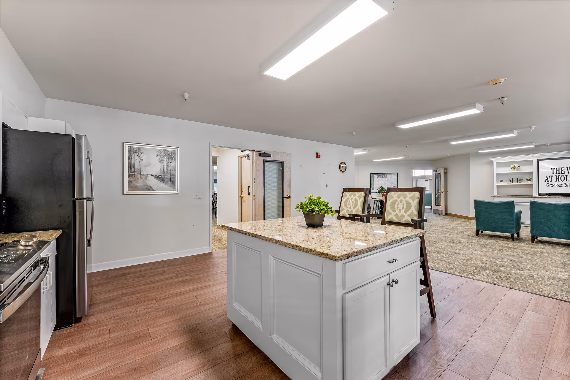 Interior view of a senior living facility showing a kitchen area with a granite countertop island and two chairs. The kitchen has a stainless steel refrigerator and stove. Beyond the kitchen is a spacious common area with carpeted flooring, teal armchairs, and a white shelving unit with a sign that reads 'The Woods at Holly Tree Gracious Retirement Living'. The walls are white, and the ceiling has rectangular fluorescent lights.